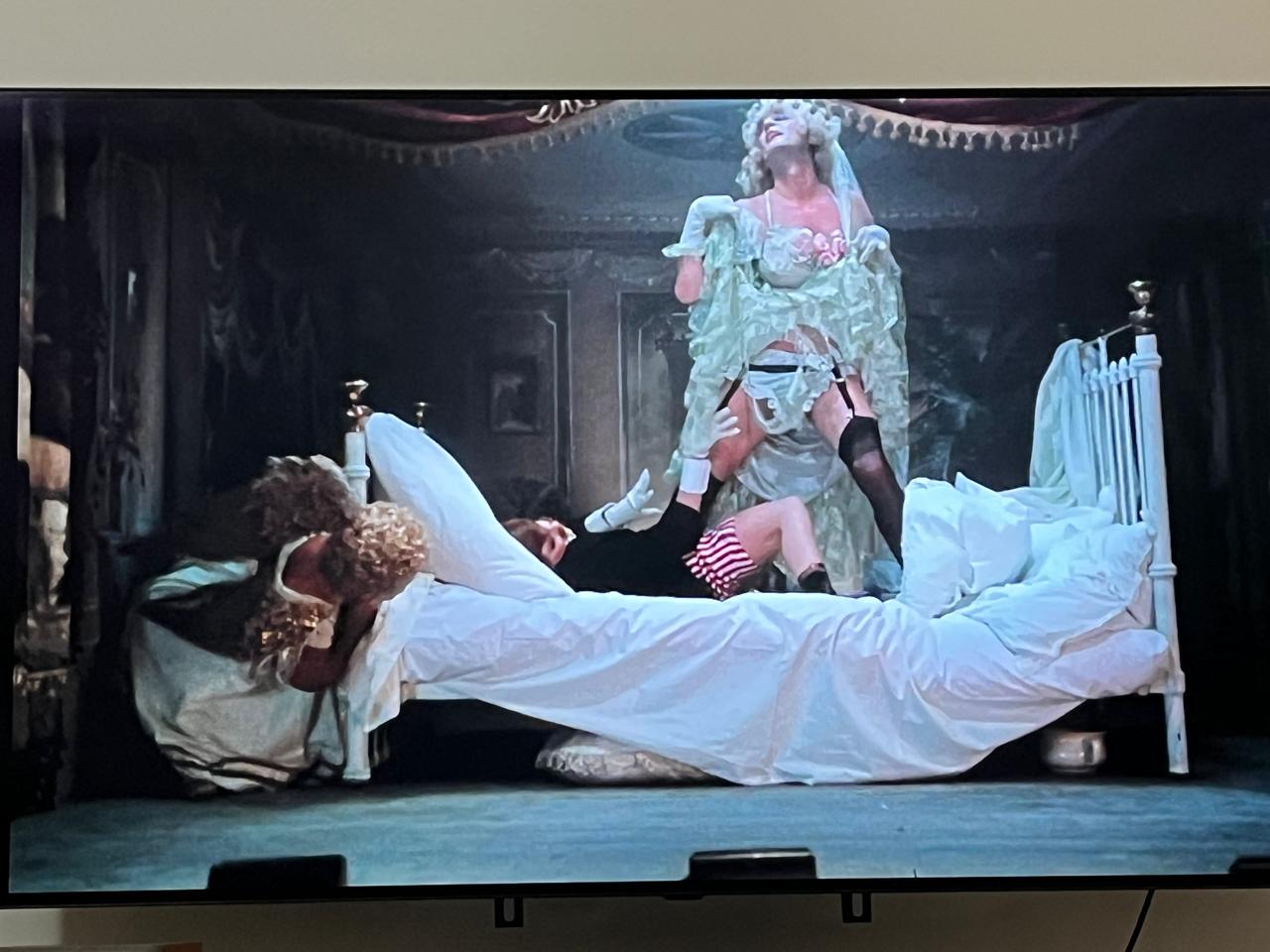 uma apresentação estilo cabaret, uma cama, um homem vestido de noiva e uma mulher vestida de noivo